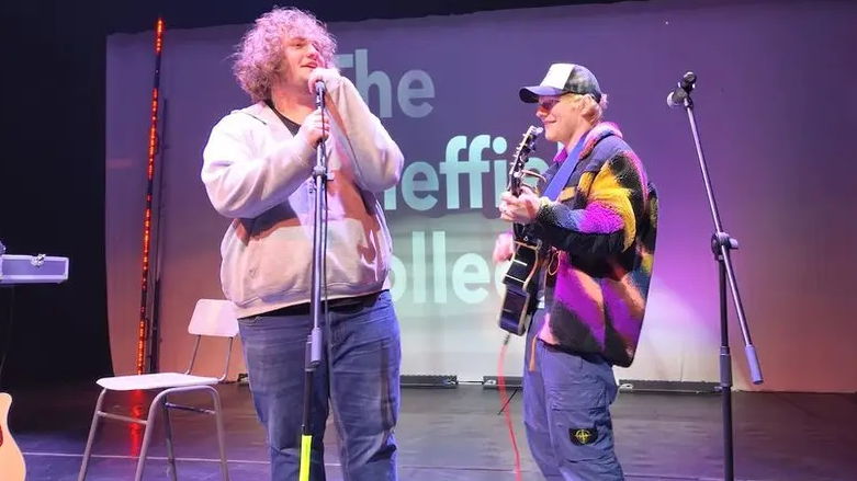 Ed Sheeran is performing with his guitar alongside a young person who's holding a microphone. Behind them, "The Sheffield College" is being projected onto a screen.