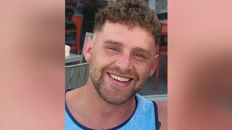 Jack Kirmond, a smiling man in his early 30s, with brown hair, stubble and a light blue vest.