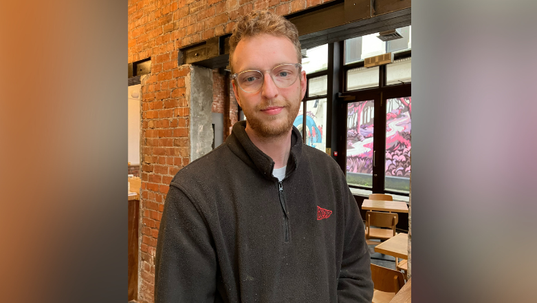 Steven is standing in his café. He wears clear glasses and has fair hair and hair on his face. He's wearing a black hoodie. The café has exposed brick walls and brown wooden tables and chairs.