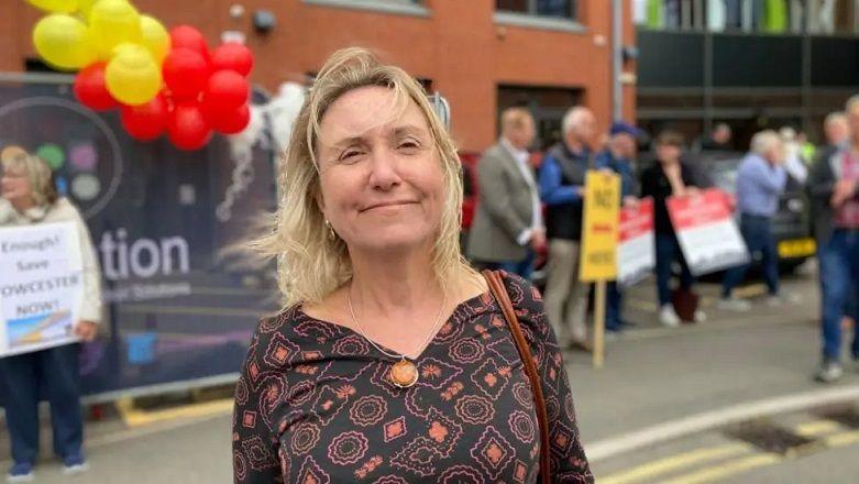 Isla Whitcroft with long blonde hair smiling and wearing a black and orange patterned dress. Protesters with placards are visible behind her, and there are several red and yellow balloons.