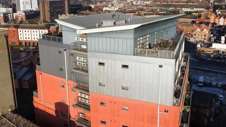 An aerial view of Cardinal Lofts which is a nine-storey block of flats. Its exterior is made up of orange and grey panels and some balconies can be seen. 
