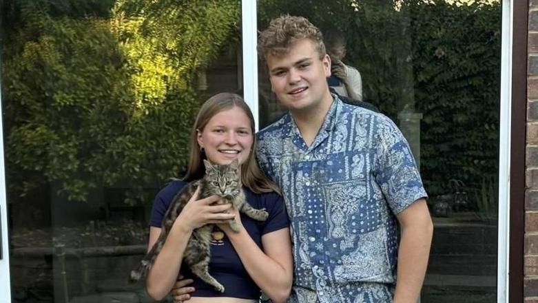 Grace Sangster and Ollie Vass standing in front of patio doors holding a young tabby cat and smiling at the camera