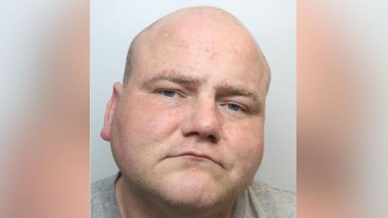 A mugshot of a bald man in his 30s with a serious expression and wearing a grey t-shirt. 