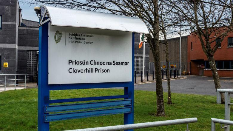 The Cloverhill Prison building is a mixture of grey and red brick. There is a patch of grass at the front with a blue and white sign indicating the name of the prison in Irish and English. 