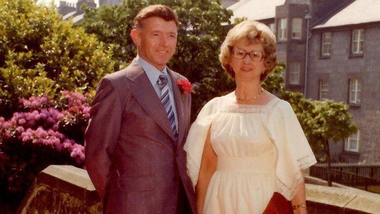 Murder victim George Murdoch and wife Jessie with flats, bushes and pink flowers behind them