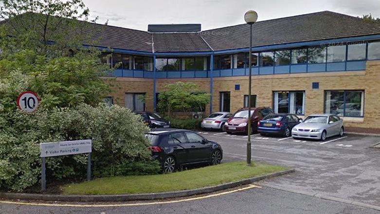 General street view image of the 2 The Pavilions in Ashton. A two-storey building, sandstone on the ground floor and with blue framed glass on the top floor and a grey roof. There is a car park to the front with greenery