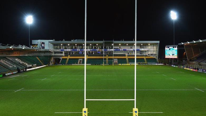 Franklin's Gardens rugby ground at night with goalposts in the foreground and stands on three sides. Two floodlights are visible left and right, and a large display screen is seen to the right.