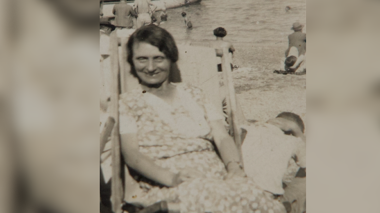 An old sepia-tone photograph of a woman at the seaside. She is wearing a floral dress and sitting in a deck chair.