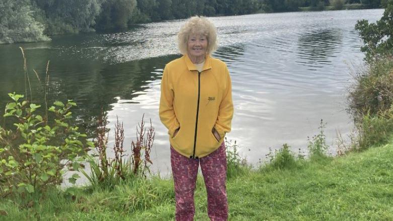 Dot Wagstaff wearing a yellow jacket, pink trousers and trainers while stood with her hands in her pockets in front of a body of water and greenery.