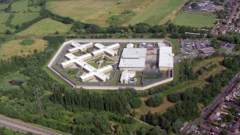 An aerial image showing HMP Bronzefield in Newbury. It is a large concrete complex with four cross shaped buildings and a large rectangular building within a surrounding perimeter wall. The entire prison is surrounded by trees and fields. 