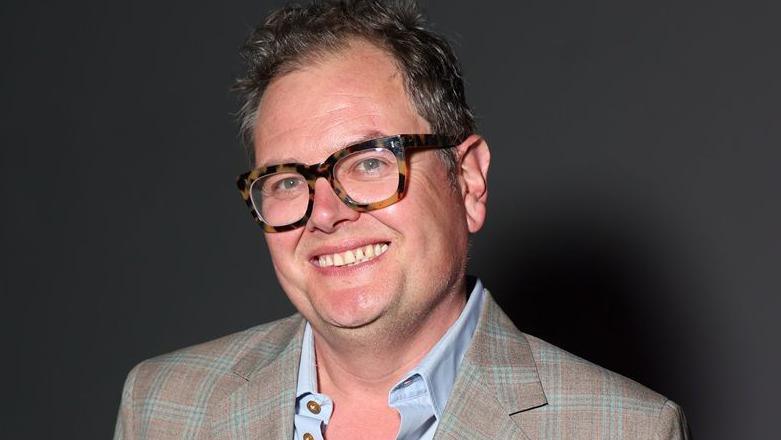 Alan Carr in a brown and light green checked suit jacket with a blue shirt underneath and brown, thick-rimmed glasses.
