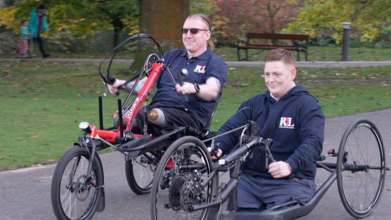 The men are both smiling as they travel on a path in a park on handcycles