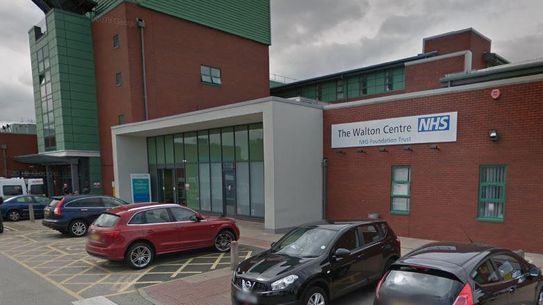 Cars parked in the car park of the Walton Centre, a redbrick health facility with a glass entrance. 