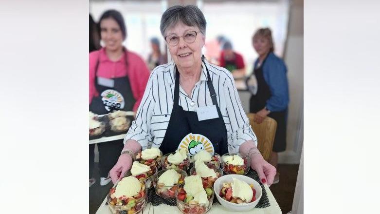 Cate is standing in the middle of the image holding a tray with food on. She has short grey hair and is wearing a white and black stiped shirt and a black apron. People are standing in the background.
