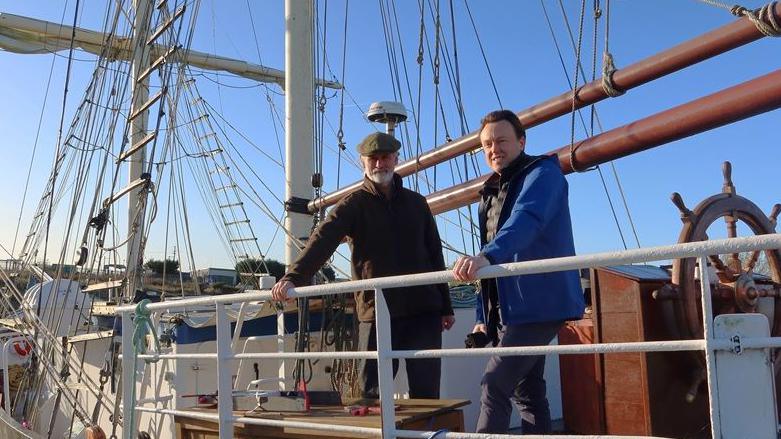 Two men on the deck of a boat on sunny day. They are wrapped up in warm coats. 