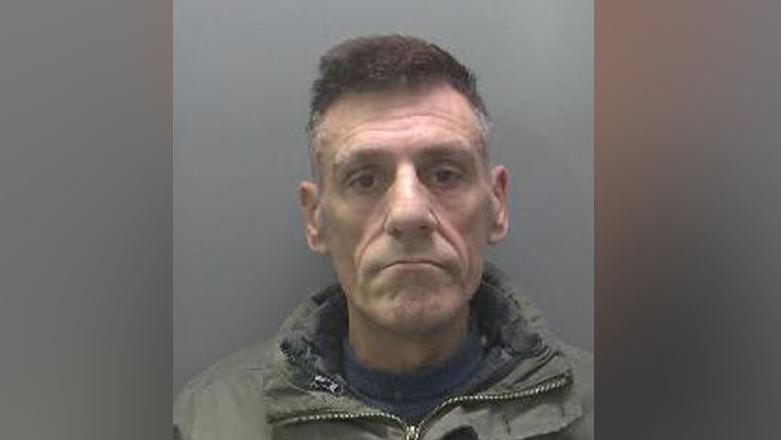 Alvaro Ferreira is looking at the camera in a police custody shot. He has short dark hair which is greying at the sides. He is clean-shaven. He is wearing a green anorak over a blue sheater