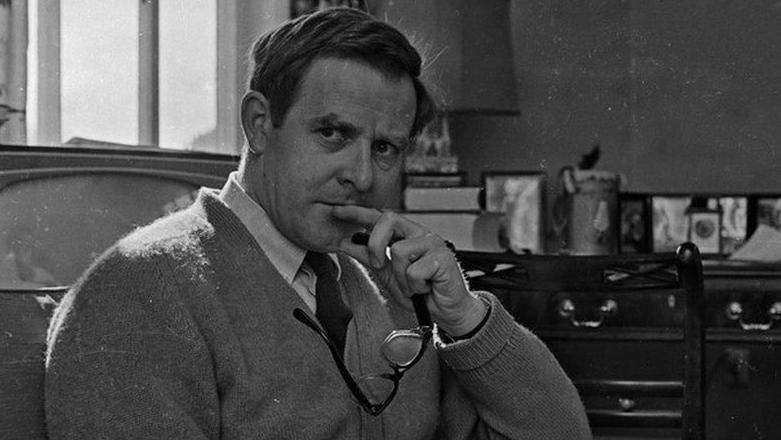 Black and white photo of Le Carré. He is looking off camera, his hand on his chin and glasses in his hand. He wears a cardigan, dark tie and a white shirt and is clean shaved with short dark hair. A chair and desk can be seen in the background. 