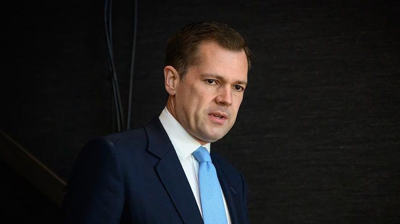 Robert Jenrick in a dark suit, white shirt and blue tie against a black background