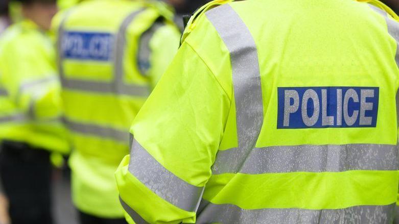 Three police officers standing outside with their backs to the camera. They are all wearing yellow fluorescent jackets which have silver stripes on the back and arms and writing which says POLICE on the back.