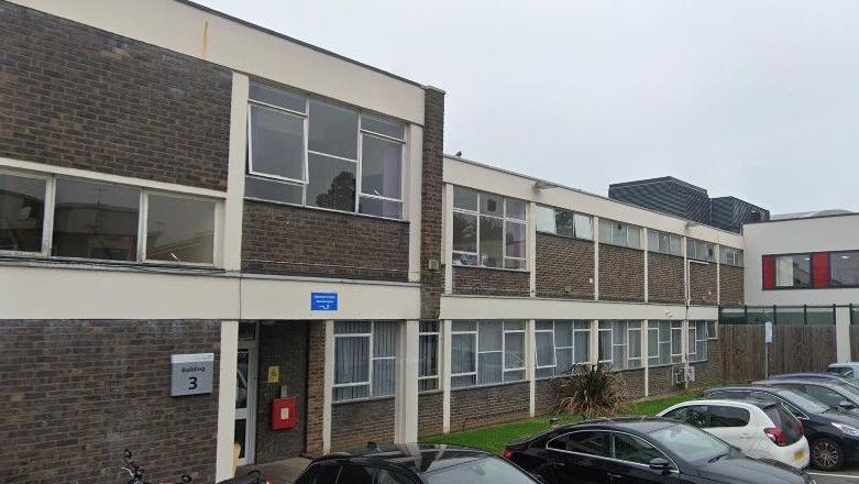 A dark brown bricked building goes across the image. The two-storey building has multiple windows and an entrance on the left. Cars are parked outside and a stretch of grass goes alongside the building.