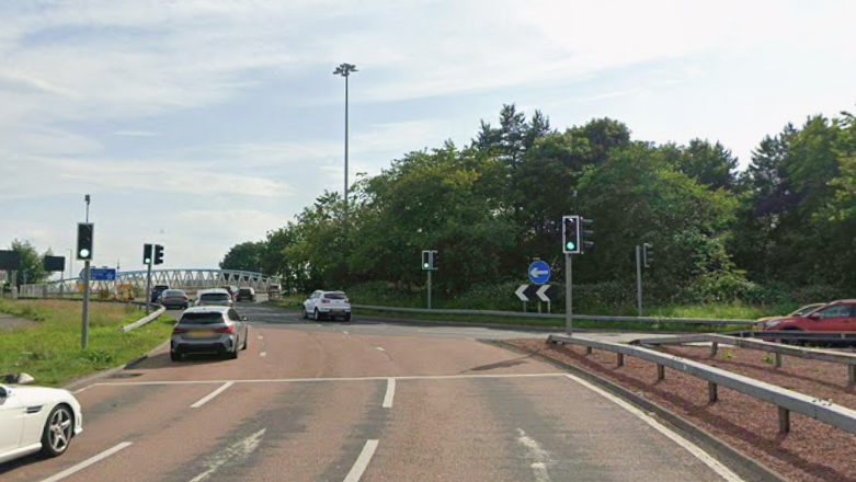Three red lanes lead up to the A92 Halbeath roundabout. The roundabout is controlled by traffic lights and the centre of the roundabout is full of trees and bushes. There are cars going up to the roundabout and also on the roundabout.