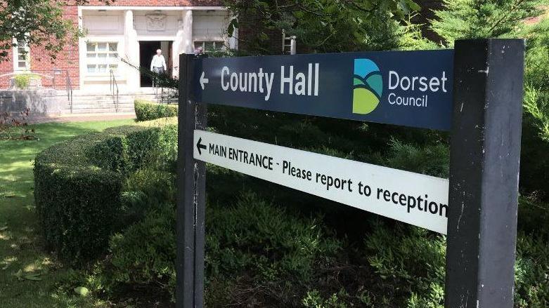 A sign near Dorset Council's headquarters points the way to the main entrance of County Hall, which is in the background behind a grassed area and hedges.