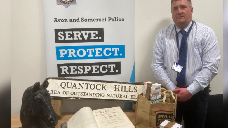 A man in a blue striped shirt, blue tie and dark trousers stands next to a table with various stolen items on it. There is a sculpture of a horse's head, a large hard-backed bible, a sign saying Quantock Hills and a hessian bag full of various packets of medication.