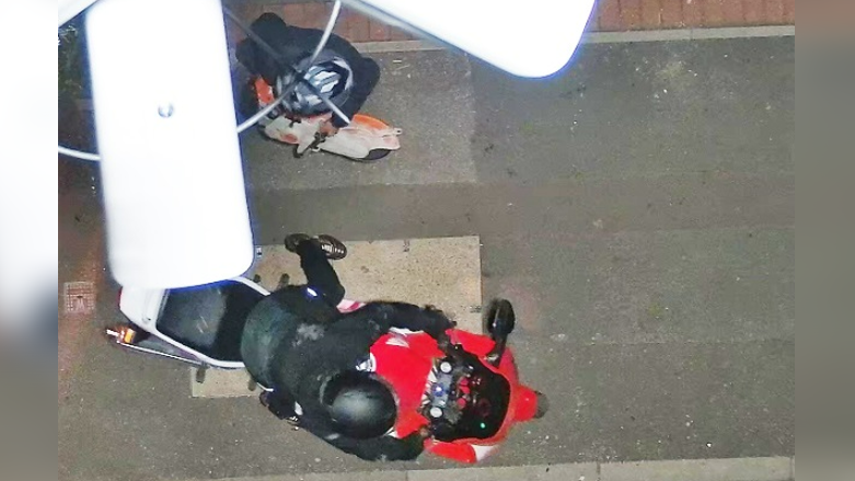 A person in black motorcycle clothing riding a red motorbike pictured from above. Another person wearing a grey striped motorcycle helmet stands to the left of the vehicle.