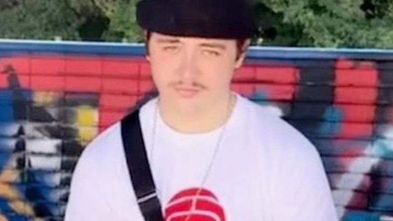 A teenager in a head and shoulders image. He is standing in front of a wall which is covered in red, black, white, blue and yellow spray paint. He is wearing a white t-shirt and a black pork pie hat, with dark brown hair at his temples and a thin pencil moustache. He has a black bag strap over his right shoulder. 