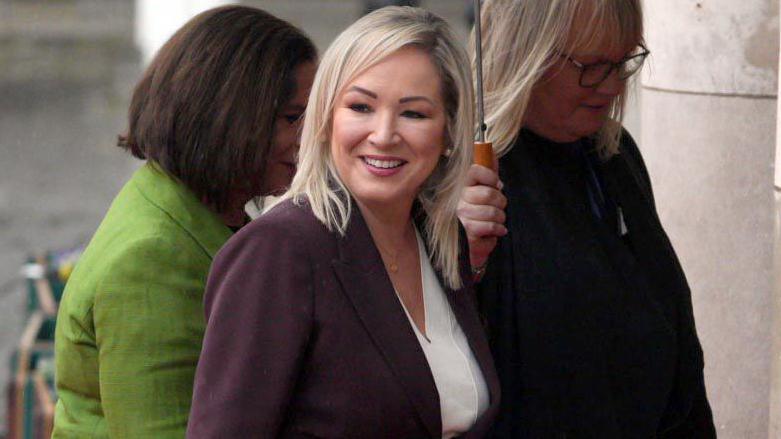 Michelle O'Neill smiling. She is wearing a purple blazer with a white top. She has blonde hair.