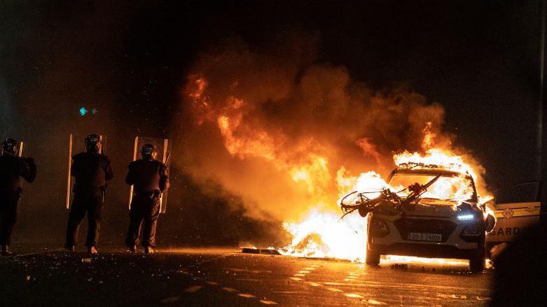 A garda police vehicle is burning in the middle of a road at night. There is a bike on the bonnet of the car. Three garda officers are standing beside the car wearing black riot gear, helmets and holding large shields that are almost body length. Photo taken on 23 November 2023. 