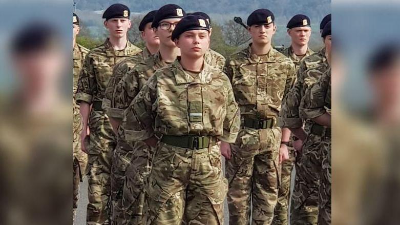 Gunner Beck wearing her camouflage military uniform and black cap. She is standing among her fellow soldiers with her arms clasped behind her back and a stern facial expression.