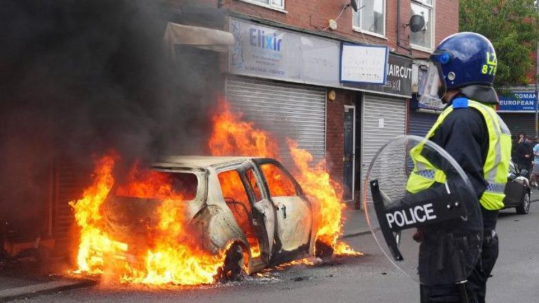 A police officer with a riot helmet and shield looks on as a car burns in front of a row of shops