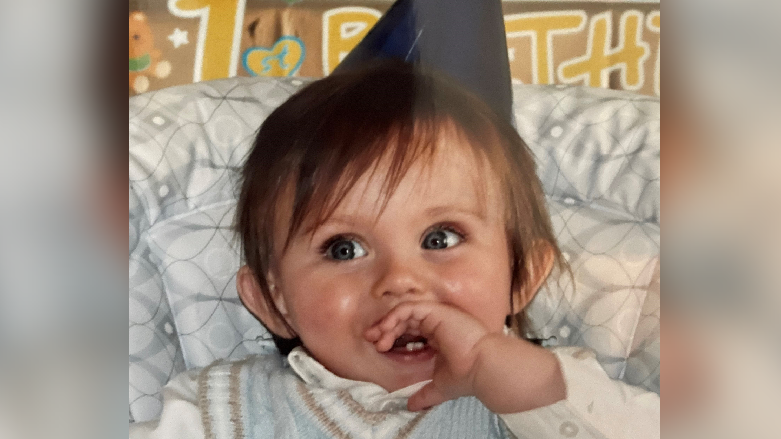 A baby smiling, he has his left hand in his mouth and is wearing a blue party hat