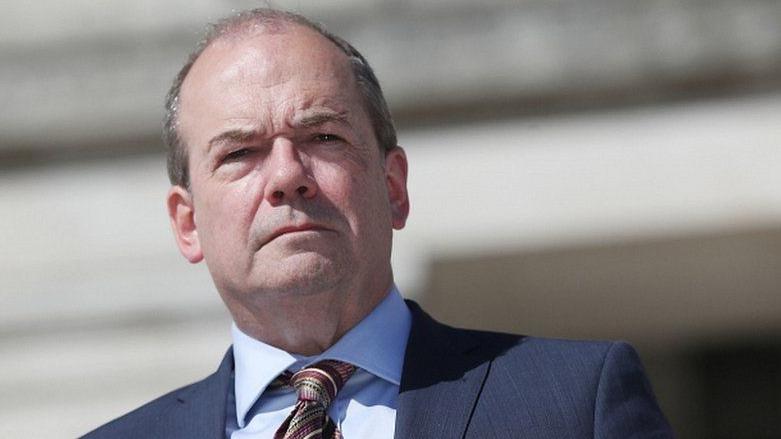 Michael McBride is looking down at the camera. He is unsmiling, with short thinning hair, and he's wearing a blue shirt and suit with a multi-coloured tie