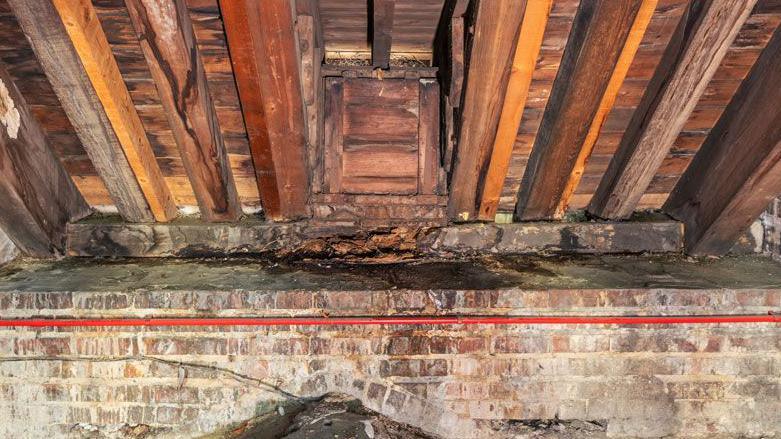 A close up of the roof timbers of the Cathedral of St John the Baptist.  The brown beams are running towards the top of the image and are in front of a plank roof lining. They are resting on a wooden beam on top of a brick built wall. The middle of the beam can be seen to be flaking away and wet with a damp sodden patch of water in front and seeping down the brick wall 