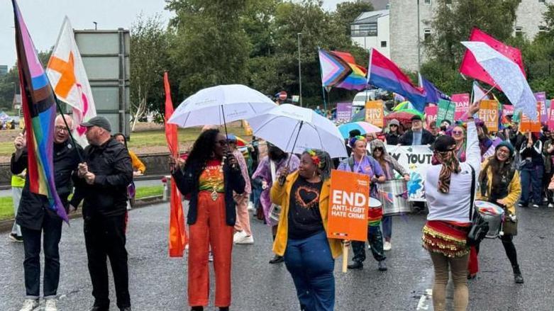 Lilian Seenoi-Barr at Foyle Pride leading crowd waving pride flags and holding umbrellas in the rain
