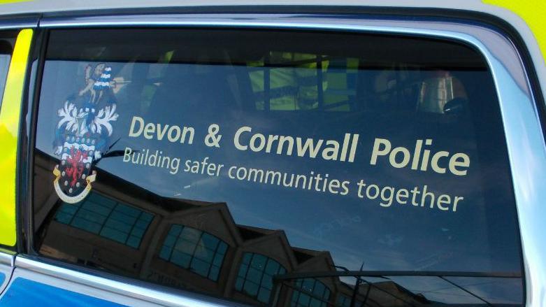 The window of a police car with the Devon & Cornwall Police logo, next to text which says Devon & Cornwall Police Building safer communities together.