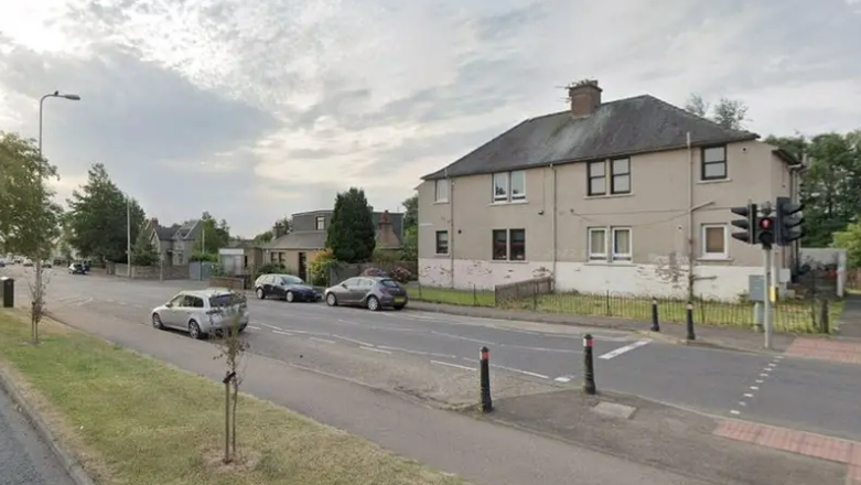 A general view of Nivensknowe Road in Loanhead. The street is empty. There is one silver car parked on the near side kerb and two dark-coloured cars parked on the far side in front of two houses. A pedestrian crossing is on the right of an image but part of it is cut off. There is a grass verge on the near side with two small sprigs attached to wooden posts.