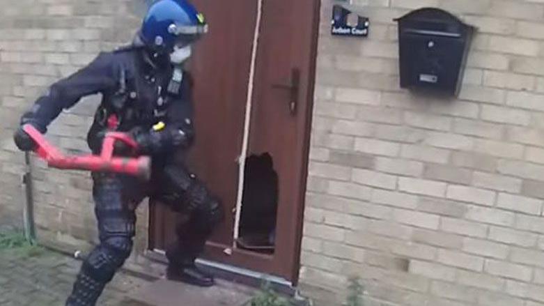 A slightly blurry bodycam footage still of a police officer swinging back a red battering ram, which is being using to bash a hole in a door. The officer is wearing black clothing and a blue motorcycle helmet and a facemask.