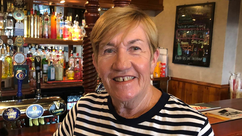 Joan Tiney standing in front of the bar at a pub while wearing a black and white striped T-shirt. She has short fair hair and is smiling.