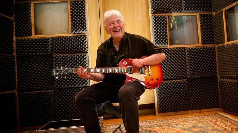 a man sits in a recording studio holding a les paul guitar. he is smiling. he is wearing a black shirt and jeans