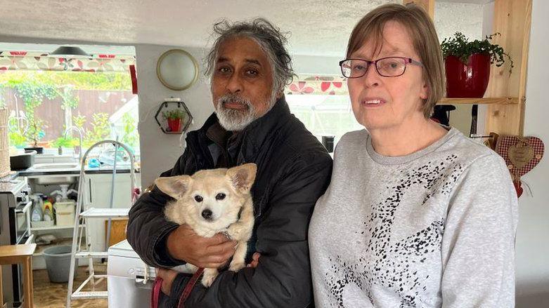 Hans and Lynn Shortt are standing in their kitchen, which has a wet floor with a bucket. Mr Shortt is carrying a small dog.