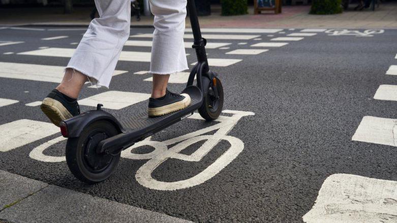 A person wearing white trousers seen from the knee down riding an e-scooter on a cycle crossing of a road.