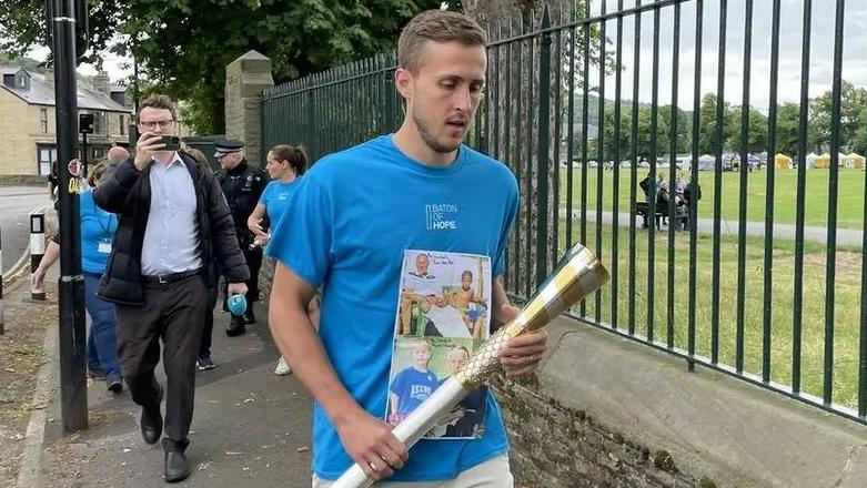 Will Vaulks waling with the baton outside Hillsborough Park. He is wearing a blue T-shirt with the words "Baton of Hope" and photographs on it. He is holding the baton.