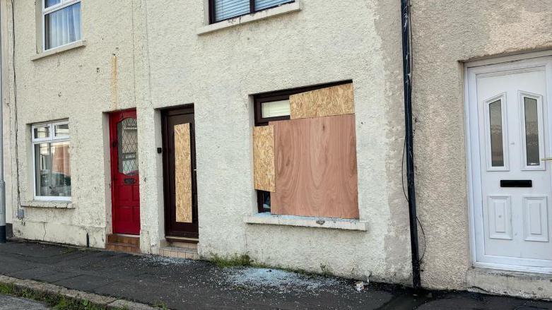 In a row of cream terraced houses, the middle one has been boarded with large wooden boards covering the front door and large front window. Broken glass lies on the footpath underneath the boarded up window.