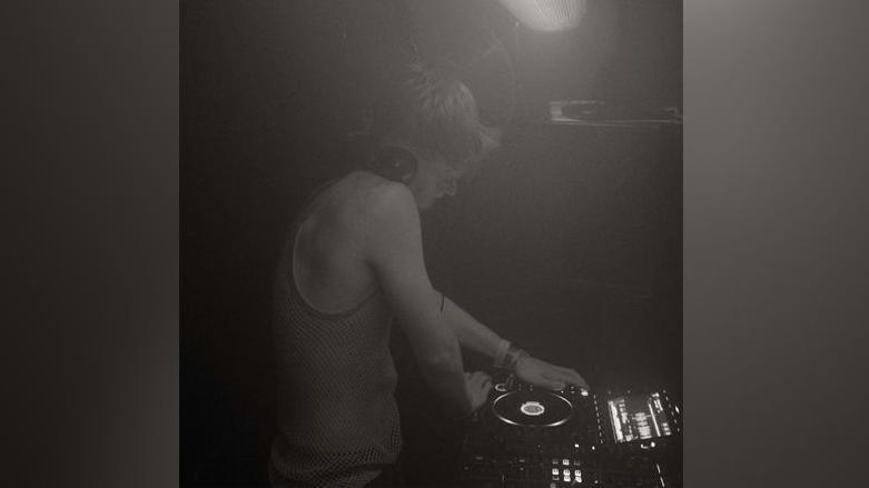 A black and white image of a young DJ playing a record. The man has short, fair hair and is wearing headphones on his head. He has a vest on.