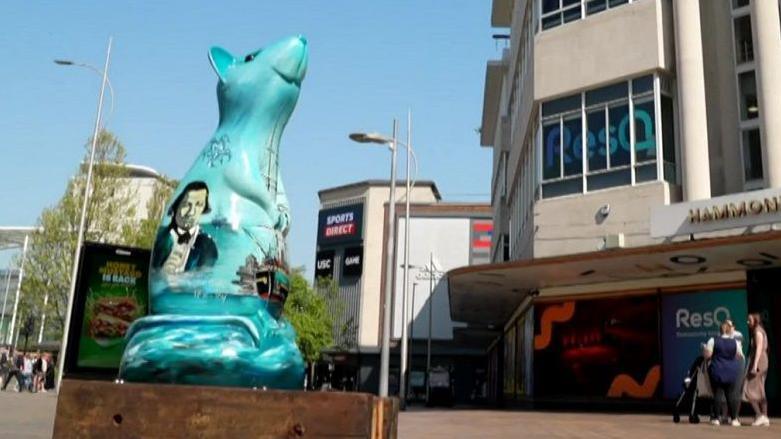 A blue rat sculpture being displayed near shops on a wooden platform
