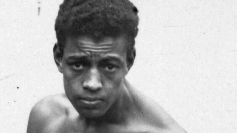 Boxer Len Johnson, shirtless and staring into the camera, stands in front of a white wall