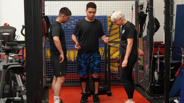 Gabriel Suarez is walking independently in a gym with two people either side of him. He is not looking at the camera but is wearing a black T-shirt and black and blue shorts.